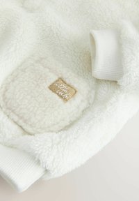 Flauschiges weißes Fleece mit einer strukturierten Oberfläche, versehen mit einem quadratischen Stoffpatch mit beigem Untergrund und weißen Nähten. Gerippte weiße Bündchen.
