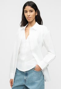 Νεαρή γυναίκα με μακριά σκοτεινά μαλλιά, που φοράει λευκό πουκάμισο με κουμπιά πάνω από λευκό μπλουζάκι και ανοιχτόχρωμα μπλε τζιν, στέκεται με το ένα χέρι στην τσέπη.