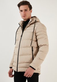 Beige pūkainis jakas ar kapuci, ar rāvējslēdzēju priekšpusē, divām sānu kabatām un melniem akcentiem uz rāvējslēdzējiem un piedurknēm. Strukturēts džemperis dizains.