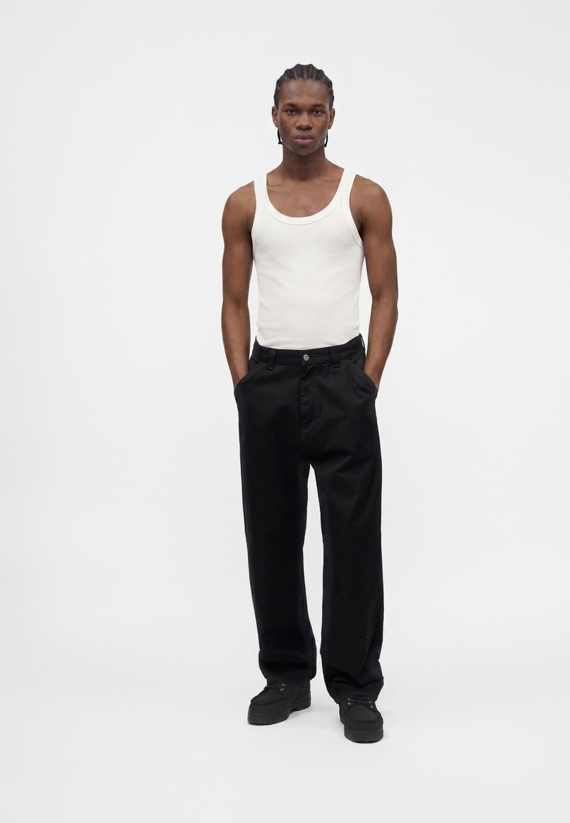 Young man with braided hair wearing a white tank top, black loose pants, and black shoes, standing with hands in pockets against a plain background.