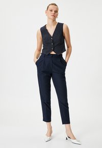 Navy-blau gestreiftes, kürzer geschnittenes Weste mit Knöpfen, kombiniert mit taillierten navy-blauen Hosen. Das Modell trägt weiße spitze Absatzschuhe. Minimalistisches Design.