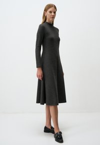 Langärmliges, eng anliegendes graues Kleid mit hohem Kragen, das am Saum ausgestellt ist. Getragen mit schwarzen Slippers, die dekorative Metallakzente aufweisen.