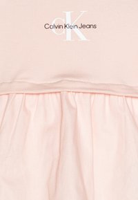 Jasnoróżowa sukienka z dopasowaną górą, marszczonym dołem i kontrastowym, ciemnym logo "Calvin Klein Jeans" na piersi. Miękki materiał.
