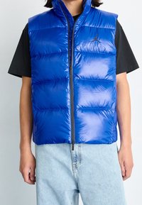 Blå puffväst med hög krage, dragkedjestängning och quiltat mönster. Tillverkad av glänsande, slätt material. Bärs över en svart T-shirt.