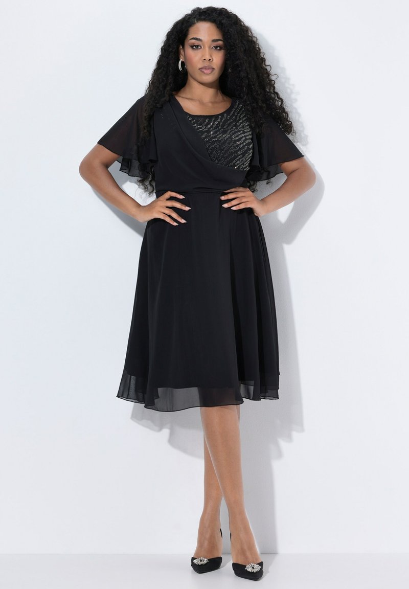 Robe noire avec des manches floues en voile, corsage ajusté avec des détails texturés. Jupe fluide tombant juste en dessous du genou, portée avec des chaussures à talons noires.