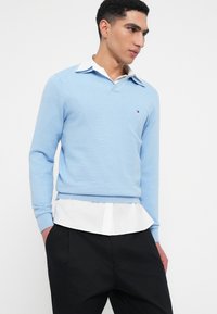 Fiatal férfi világoskék bordázott pulóverben fehér inggel és fekete nadrággal, kezét a zsebébe dugva, jobbra nézve fehér háttér előtt.