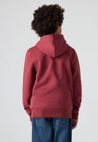 Sudadera de color burdeos con una gran capucha, puños y dobladillo acanalados. Textura de tela suave, combinada con jeans de mezclilla azul. Diseño sencillo, sin logotipos visibles.