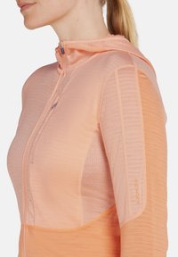 Veste à capuche zippée en orange clair, dotée d'un tissu texturé, de panneaux lissés contrastants et d'accents en maille sur les côtés.