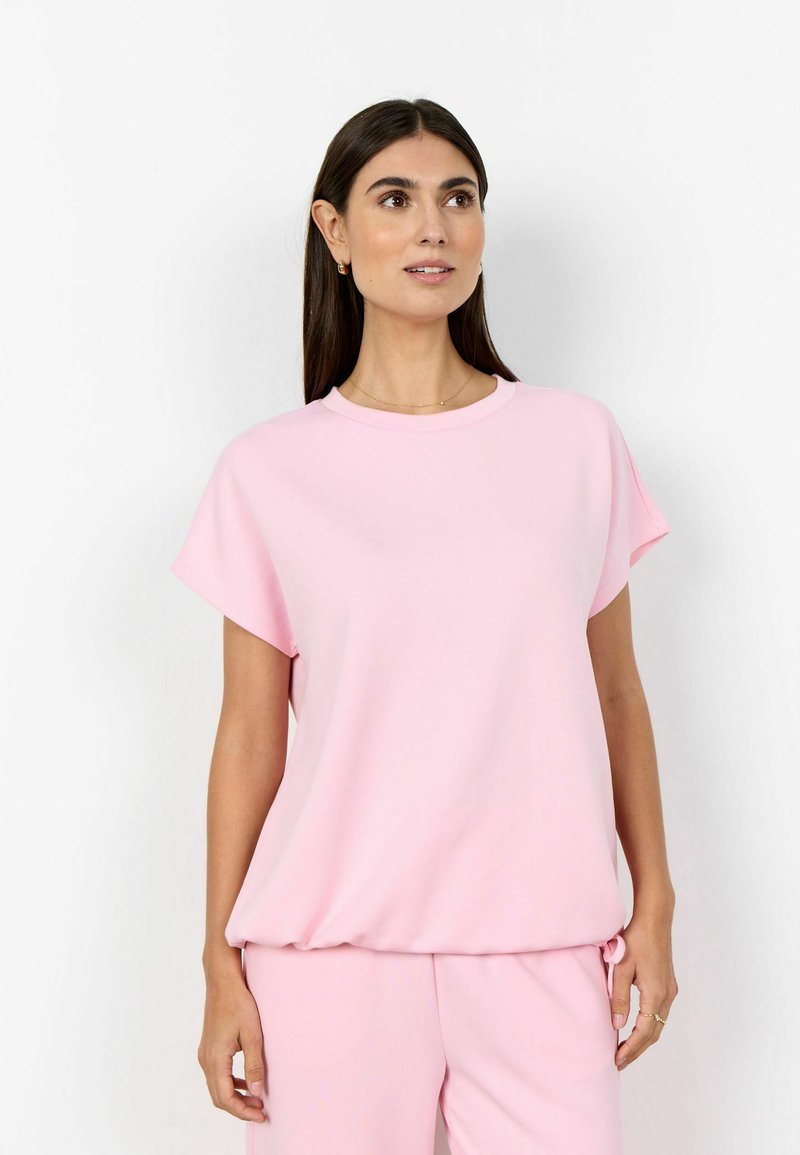 Woman with long dark hair wearing a loose light pink short-sleeve top and matching pants, standing against a plain white background.