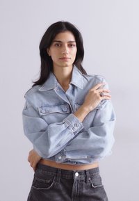 Jachetă din denim bleu deschis cu croială supradimensionată, cu închidere prin butoane și buzunare la piept, purtată peste blugi negri cu talie înaltă. Textură netedă.