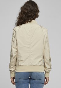 Beige bomber jacket with a smooth, shiny texture, ribbed cuffs, and a fitted waistband. Includes side mesh panels and streamlined seams.