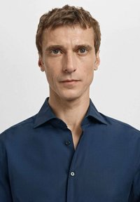 Granatowa koszula z kołnierzem na guziki, subtelnym połyskiem i dwoma guzikami z przodu. Tkanina wydaje się gładka, bez wzorów ani faktur.