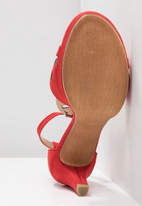 Chaussure à talons hauts en daim rouge avec un bout en amande, une délicate bride de cheville et une semelle en caoutchouc lisse de couleur brun clair avec de fines rainures.