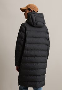 Svart dunjakke med horisontal quilting, som har høy krage og avtagbar hette. Modellen har også på seg en tan cap og blå jeans.