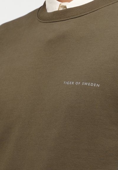 Φόρμα ελιάς με στρογγυλή λαιμόκοψη, κατασκευασμένη από λείο βαμβακερό ύφασμα. Διαθέτει μικρό λευκό λογότυπο που λέει "TIGER OF SWEDEN."