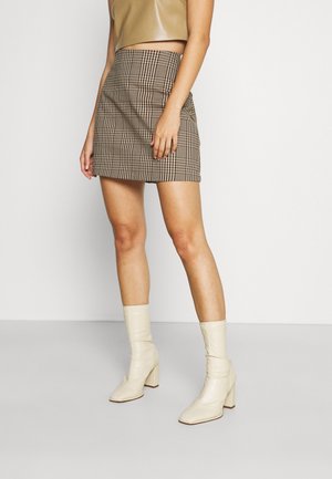 Femme portant un crop top beige, une mini-jupe à carreaux marron et des bottines crème à talons hauts, debout devant un fond uni.