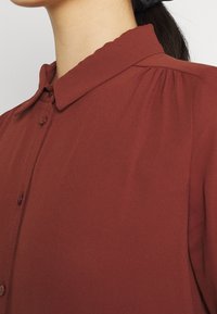 Chemise boutonnée bordeaux avec col, détails plissés aux épaules et texture lisse. Présente de petits boutons et un design épuré.