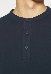 Närbild av en person som bär en mörk marinblå henley-tröja med tre svarta knappar och rund halsringning.