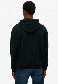 Sudadera negra con un corte relajado, una capucha grande y puños acanalados. Se lleva con vaqueros de mezclilla azul, mostrando un diseño minimalista.