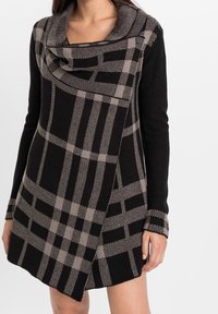 Abito lavorato a maglia con scollatura a barchetta, caratterizzato da un motivo a quadri nero e beige, maniche lunghe e un orlo leggermente asimmetrico.