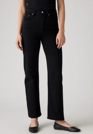 Femme portant un jean noir taille haute, coupe droite, des ballerines noires et un top noir sans manches sur un fond uni.