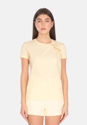 T-shirt giallo chiaro a maniche corte in tessuto morbido con dettaglio plissettato e un accento dorato sulla spalla. Orlo corto, design aderente.