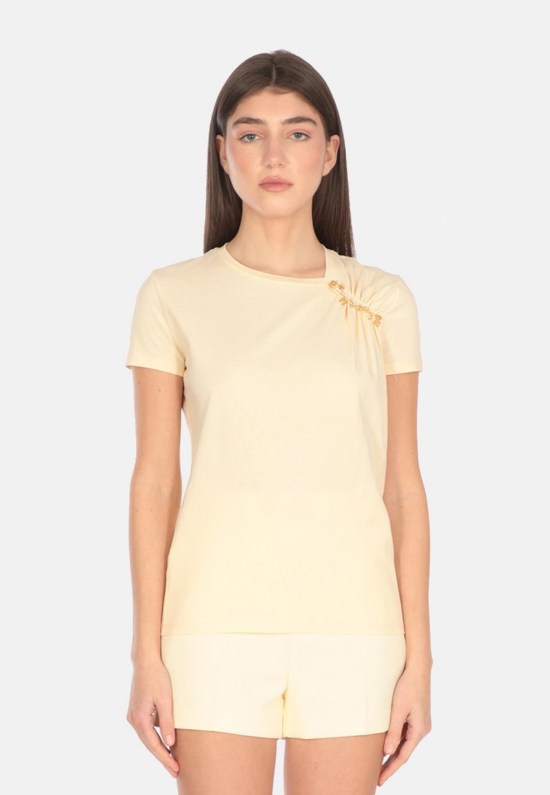 T-shirt giallo chiaro a maniche corte in tessuto morbido con dettaglio plissettato e un accento dorato sulla spalla. Orlo corto, design aderente.
