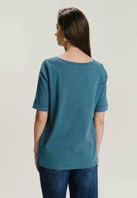 Turkusowa bawełniana koszulka z szerokim dekoltem i krótkimi podwiniętymi rękawami, z subtelnym wzorem z tyłu. Zestawiona z niebieskimi dżinsami.