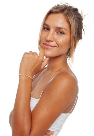 Bracelet avec un charm en forme de cœur doré et de petites perles dans des couleurs pastel. Le modèle touche délicatement son menton, portant un débardeur blanc et ayant des cheveux mi-longs.
