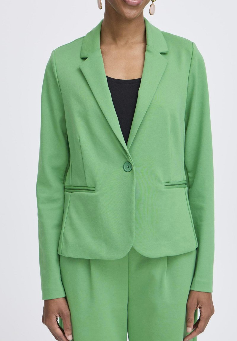 Vrouw draagt een felgroene blazer en bijpassende broek over een zwarte top, staand tegen een effen lichte achtergrond.