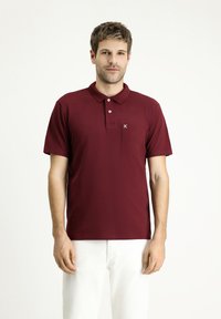 Polo bordeaux con patta a due bottoni, maniche corte e taschino sul petto che presenta un piccolo logo. Textura liscia, abbinato a pantaloni bianchi.