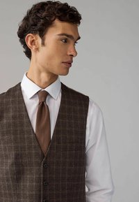 Brązowa kamizelka w kratę noszona na białej koszuli, zestawiona z teksturowanym brązowym krawatem. Posiada slim fit i zapięcie na pięć guzików.