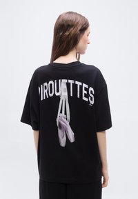 Νεαρή γυναίκα που κοιτάζει προς τα πίσω φορώντας μια μαύρη, φαρδιά μπλούζα με το γράμμα «PIROUETTES» και μια απεικόνιση πουλιών μπαλέτου στην πλάτη.