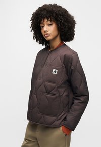 Καφέ επενδυμένο μπουφάν με καπιτονέ σχέδιο, μπροστινή τσέπη και κούμπωμα με κουμπιά. Διαθέτει έντονο πορτοκαλί γιακά και λεία υφή υφάσματος.