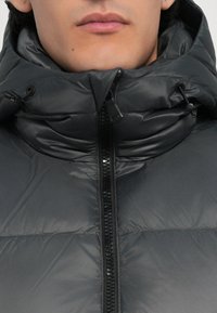 Hallhalli halljahoidjaga jakk kõrge kaelusega, tõmbluku kinnitusega ja reguleeritava paelaga. Läikiv tekstuur, õmmeldud paneelid ja musta tõmbluku detail.