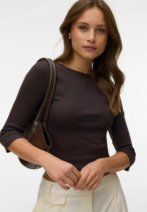 Młoda kobieta o brązowych włosach, ubrana w dopasowaną ciemnobrązową bluzkę i kremowe spodnie, trzymająca brązową torebkę na ramieniu na jednolitym tle.