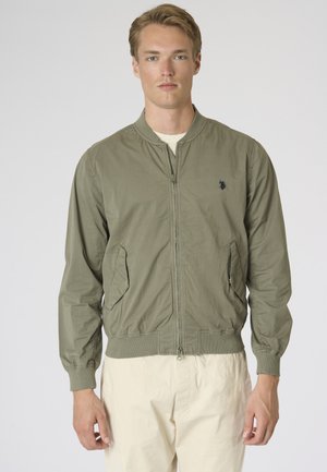 Giovane uomo che indossa una giacca bomber verde oliva chiaro con cerniera e pantaloni color crema, in piedi davanti a uno sfondo chiaro e uniforme.