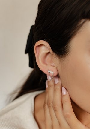 Silberne Ohrringe mit drei weißen Perlen, die durch eine zarte Kette verbunden sind, vor einem sanft verschwommenen Hintergrund. Eine Hand mit heller Hautfarbe berührt das Ohr.