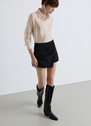 Persona che indossa una camicia beige con bottoni, minigonna nera e stivali neri alti fino al ginocchio, in piedi su un pavimento grigio chiaro uniforme.
