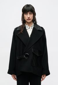 Čierny veľký kabát s klopovým golierom, koženými detailmi, veľkým opaskom so sponou v tvare kruhu a lesklým detailom na vrecku, vrstvený cez košeľu s bodkami.
