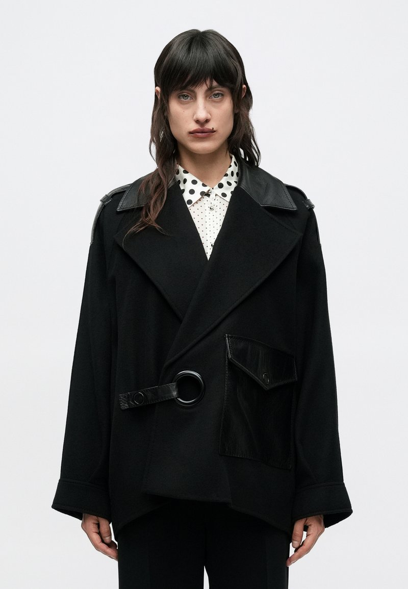 Čierny veľký kabát s klopovým golierom, koženými detailmi, veľkým opaskom so sponou v tvare kruhu a lesklým detailom na vrecku, vrstvený cez košeľu s bodkami.