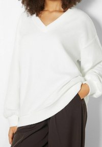 Maglione bianco a scollo a V con maniche lunghe e leggermente a sbuffo, dal taglio rilassato. Tessuto morbido, indossato sopra pantaloni marroni scuri.