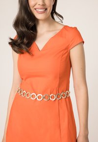 Oranje jurk met een V-hals, korte mouwen en een getailleerde taille. Geaccentueerd met een gouden kettingriem met ronde, gelinkte accenten.