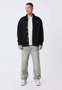 Schwarzes Hemd mit strukturiertem Stoff über einem hellen T-Shirt, kombiniert mit hellen Jeanshosen und weißen Sneakers.