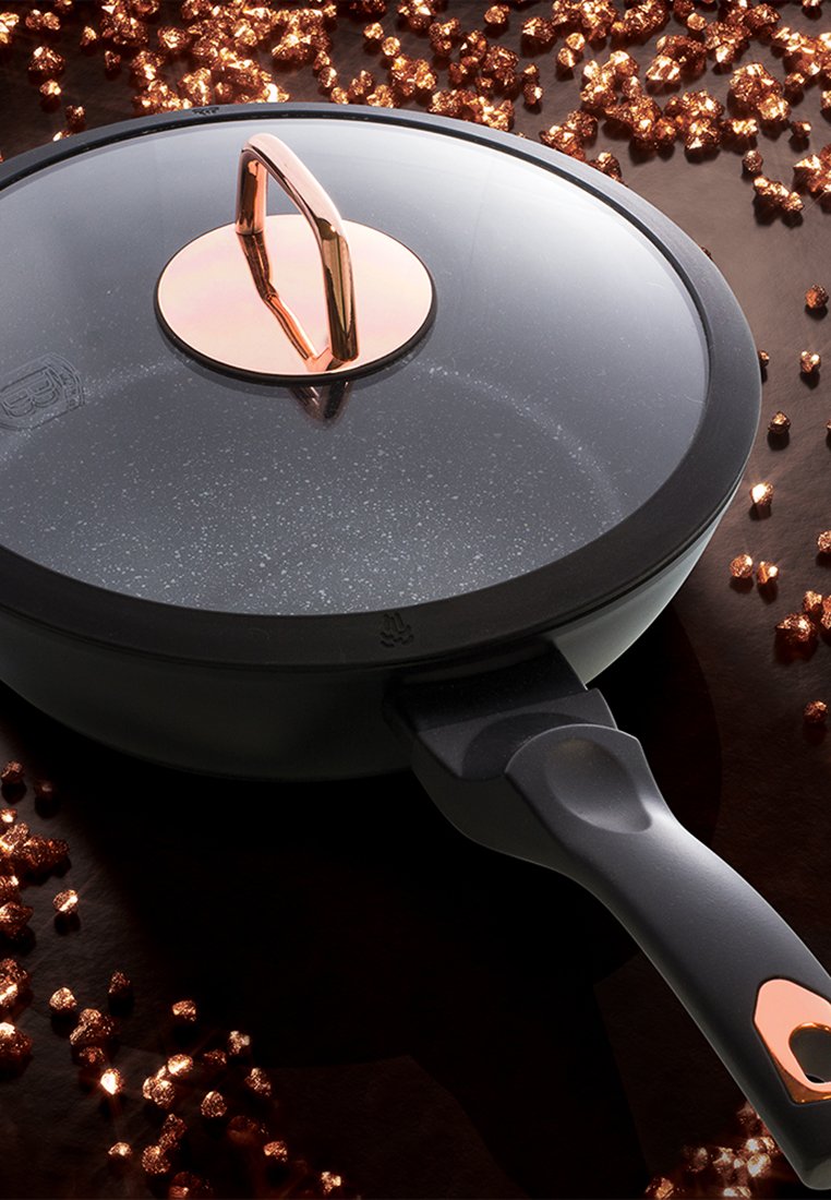 Sartén antiadherente con superficie negra moteada, tapa de vidrio y mango de cobre. Rodeada de pequeñas decoraciones metálicas de cobre.