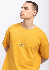 Geel katoenen t-shirt met korte mouwen, met een abstract grafisch ontwerp aan de voorkant in donkergrijs en oranje. Ronde halslijn, ontspannen pasvorm.