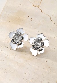 Silver-tone floral stud earrings with five reflective petals and textured centers on a beige stone surface.