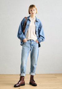 Veste en denim, polo beige, jeans bleu clair avec ourlets retroussés, et bottes marron à lacets. Comprend un sac à épaule et des textures lisses.