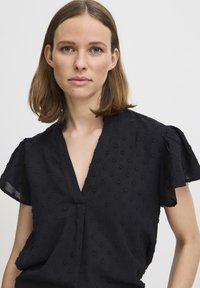 Blusa nera con scollatura a V, maniche corte a sbuffo e motivo testurizzato con piccole macchie in rilievo su tutto il tessuto.