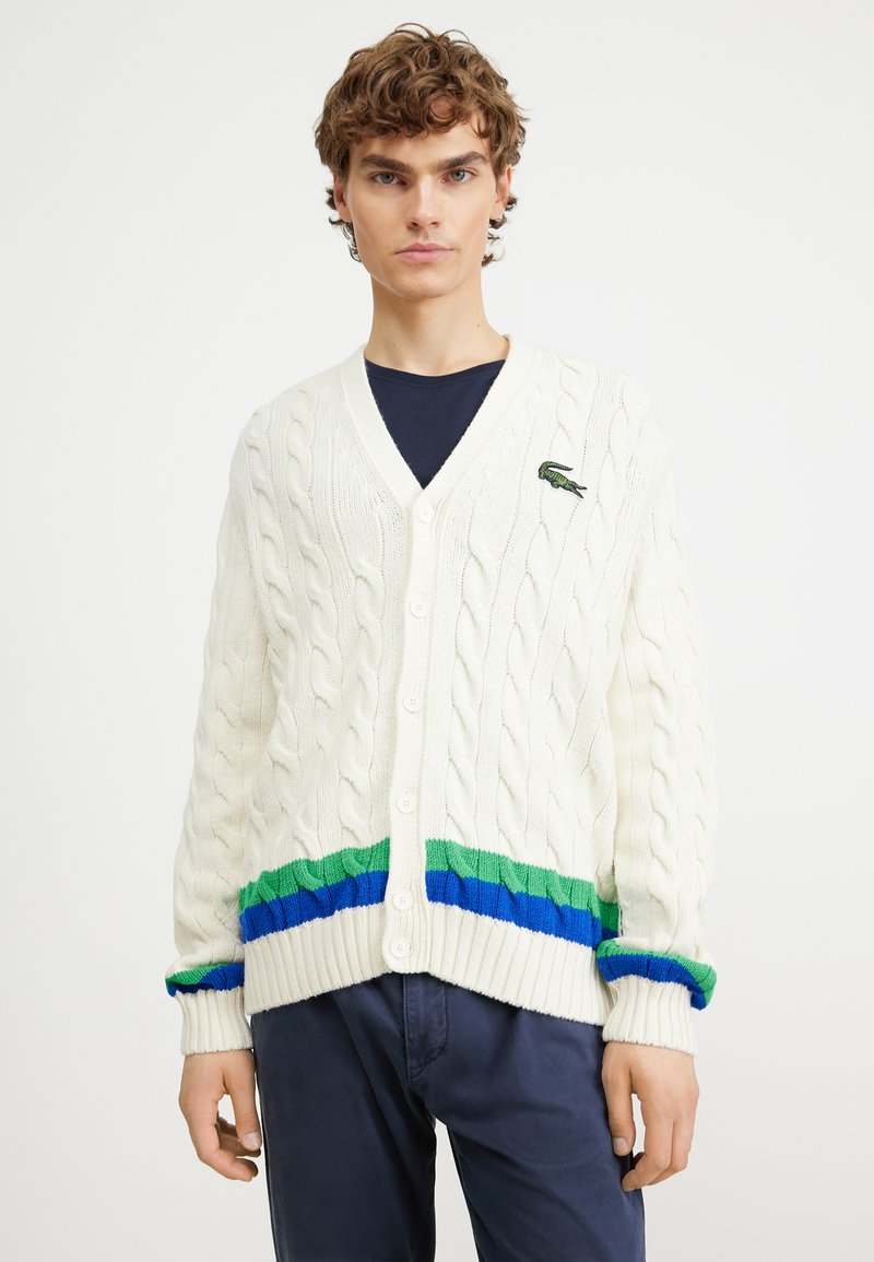 Cardigan blanc en tricot avec un motif en cable, présentant un ourlet et des poignets rayés vert et bleu, complété par un petit logo brodé vert.
